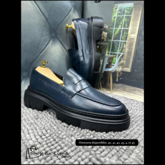 Mocassins homme bleu nuit à semelle épaisse crantée posés sur tapis gris avec embauchoirs dorés – chaussures modernes élégantes sans lacets